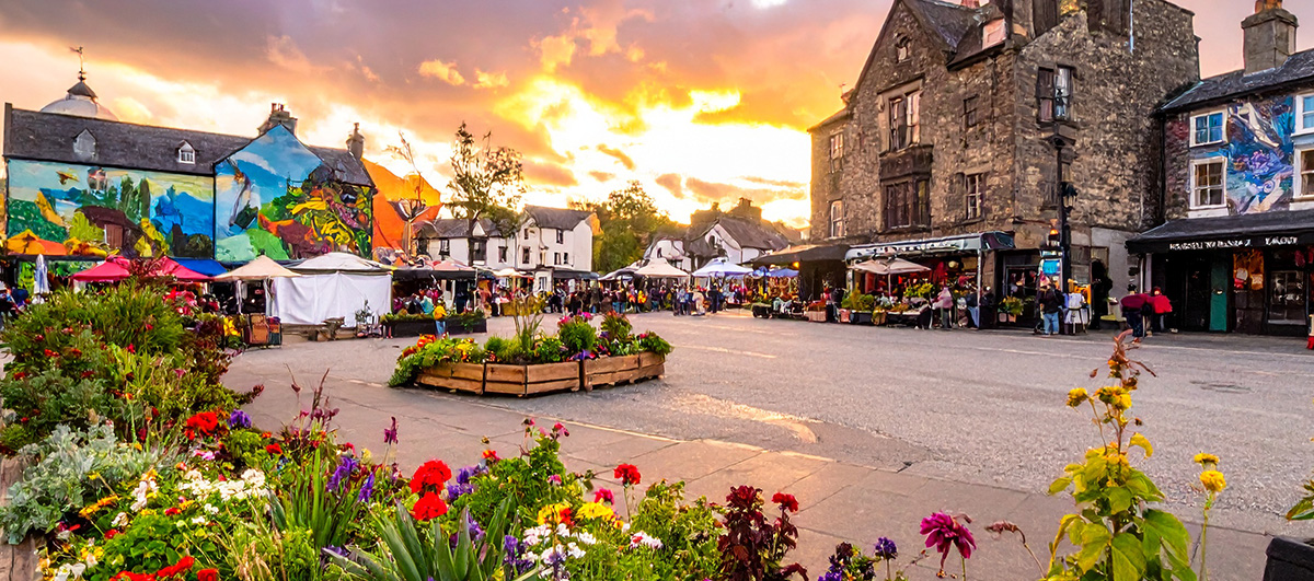 A vibrant Welsh UK town square being revitalised by volunteers, with murals, greenery, and local markets, golden hour lighting, wide-angle lens, hopeful atmosphere. 
