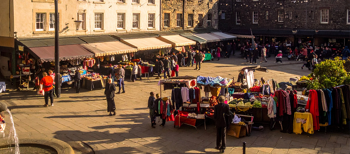 A vibrant open-air market in a historic town square, diverse stalls overflowing with colourful goods, people exchanging smiles and handshakes, children playing near a fountain, warm golden-hour sunlight casting long shadows.