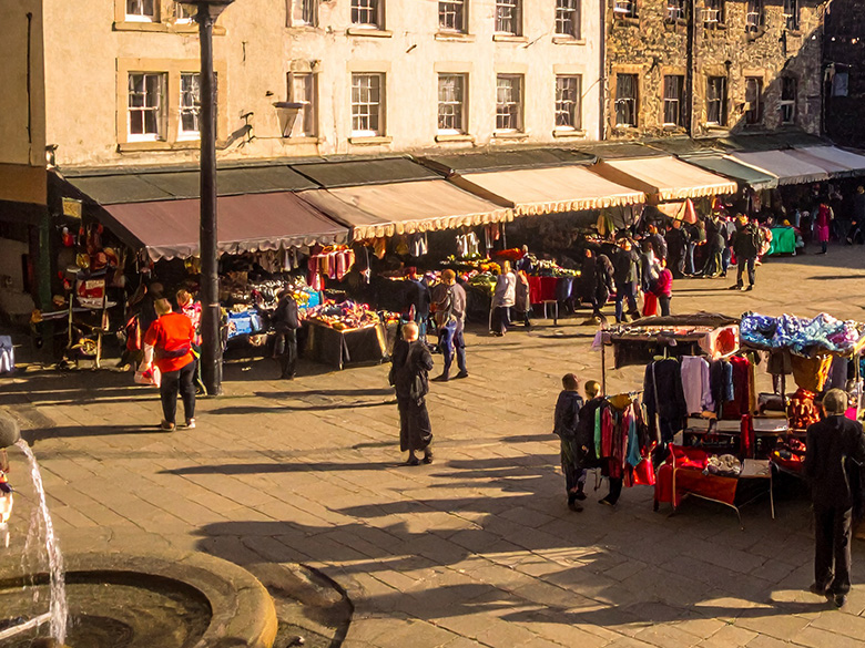 A vibrant open-air market in a historic town square, diverse stalls overflowing with colourful goods, people exchanging smiles and handshakes, children playing near a fountain, warm golden-hour sunlight casting long shadows.
