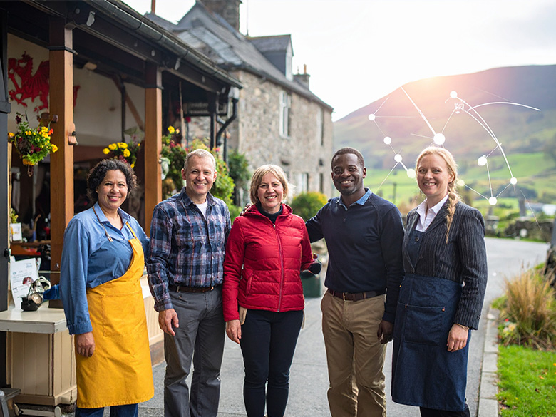 A diverse group of small business owners and entrepreneurs gathered outside a traditional Welsh shop, smiling and collaborating.